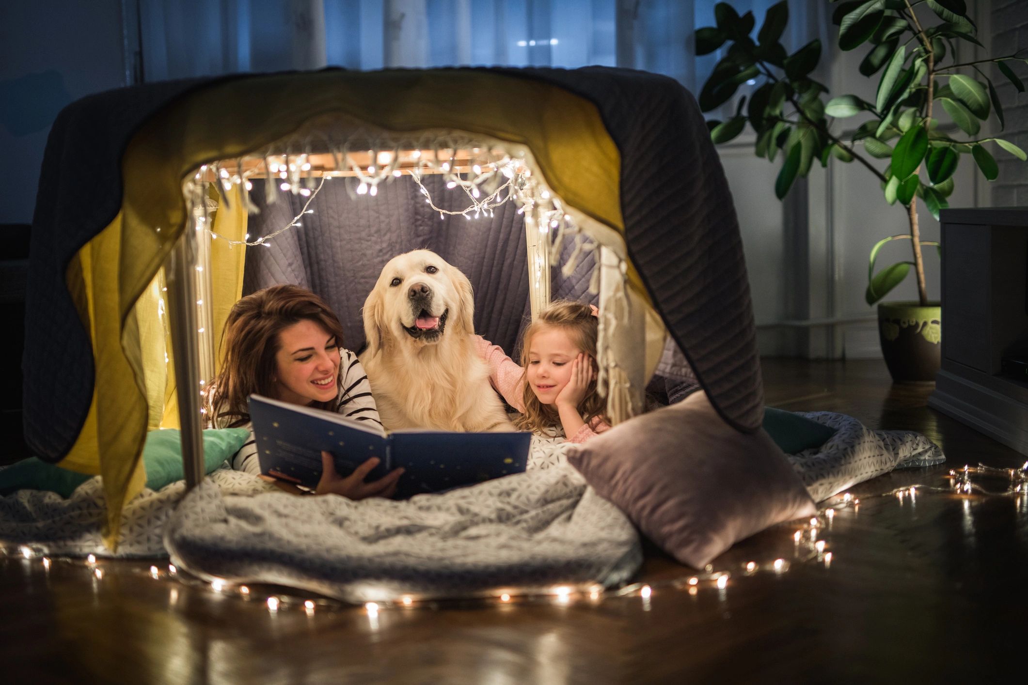 Two girls and a dog reading a book together under cozy fairy lights.